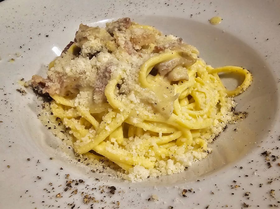 Piatto fondo bianco con pasta lunga mantecata, guanciale a pezzi, pecorino grattugiato in abbondanza e pepe nero macinato grosso sparso anche sul bord
