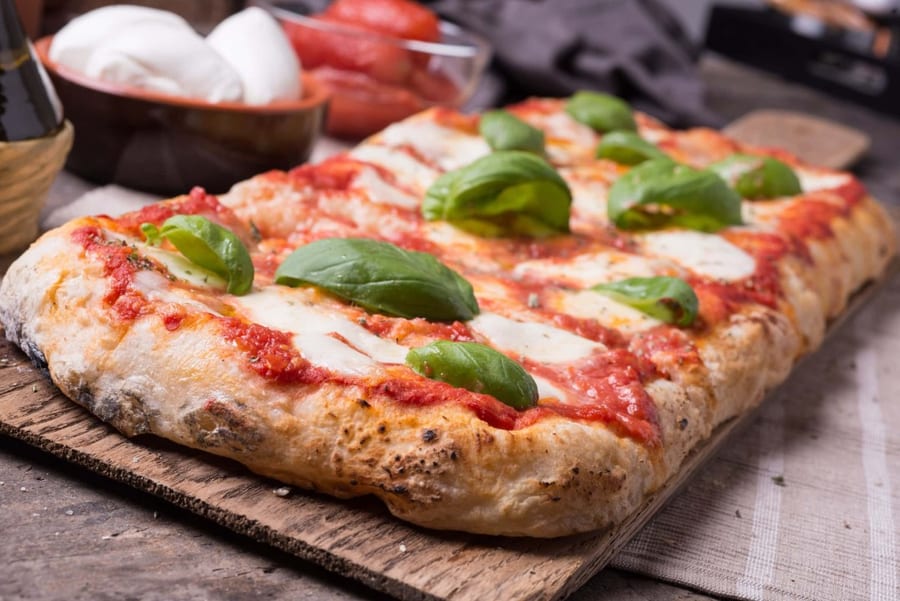 Pizza margherita su tagliere di legno rustico, con mozzarella, pomodoro e foglie di basilico fresco, ciotola di mozzarella e pomodori sfocati sullo sf
