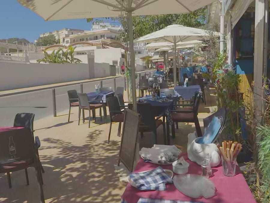 La terrazza apparecchiata vista dal tavolo: tovaglie a quadretti blu e rosse, piatti a forma di conchiglia, grissini nel bicchiere, ombrelloni aperti