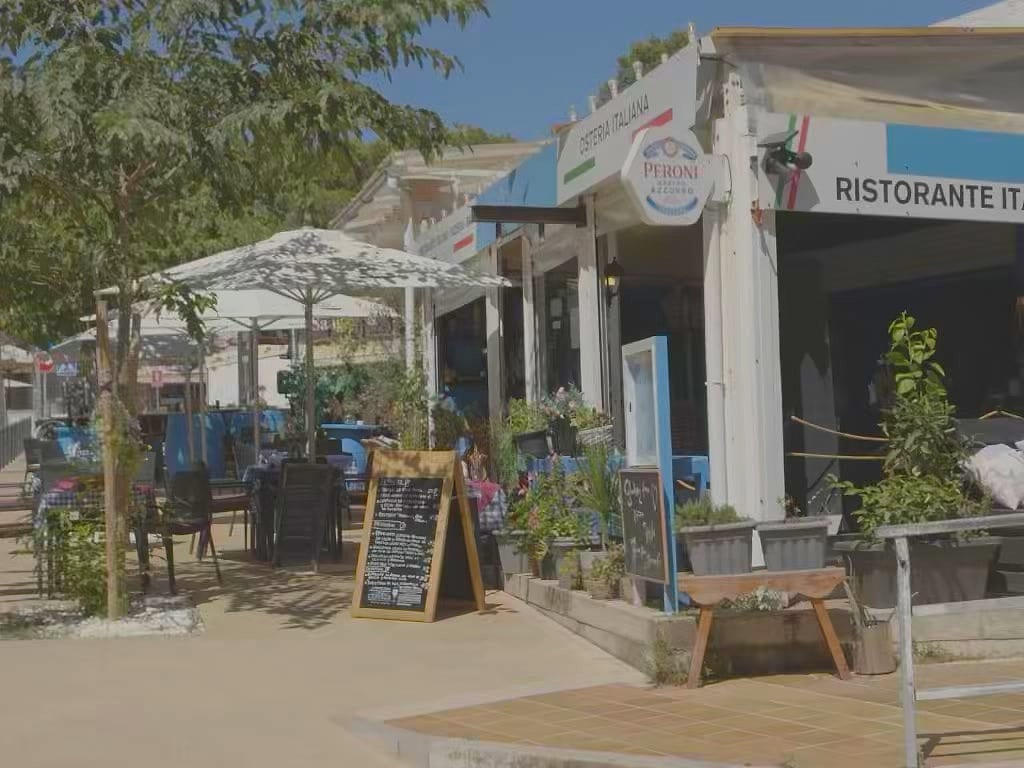 L esterno del locale dalla strada: insegna OSTERIA ITALIANA e RISTORANTE ben leggibili, ombrelloni bianchi, lavagne dei menu, fioriere e tavoli con to